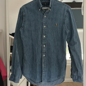 Blå jeansskjorta från Ralph Lauren - Säljer en klassisk blå jeansskjorta från Ralph Lauren modell slim fit med knappar hela vägen och broderad logga på bröstet. Skjortan har lång ärm, button down-krage och är gjord i mjukt jeanstyg. Perfekt för en avslappnad och stilren look.