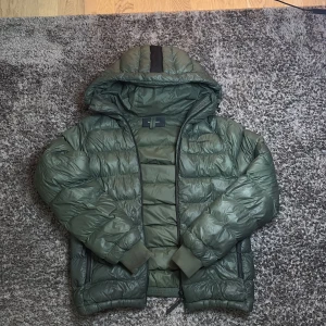 Grön dunjacka Peak Performance S - Snygg grön dunjacka från Peak Performance i storlek S. Jackan har huva, dragkedja framtill och två sidofickor med dragkedja. Puffig modell med ribbade muddar och diskret logga på bröstet. Perfekt för kalla dagar och riktigt skön att bära.