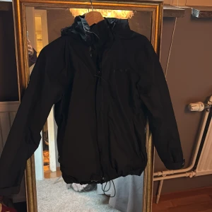 Svart vindjacka från Everest, strl 34 - Svart vindjacka från Everest med huva och dragsko nertill. Jackan har dragkedja framtill och är perfekt för blåsiga dagar. Enkel och stilren design som funkar till det mesta. Materialet är syntet och jackan har långa ärmar.