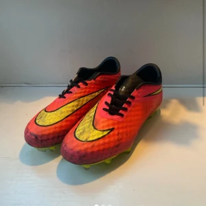 Nike Hypervenom fotbollsskor rosa/gul - Säljer ett par Nike Hypervenom fotbollsskor i en riktigt snygg rosa färg med stora gula swoosh-loggor och svarta detaljer. Skorna har snörning och platt sula med dobbar för gräsplan. Perfekta för dig som vill sticka ut på planen!