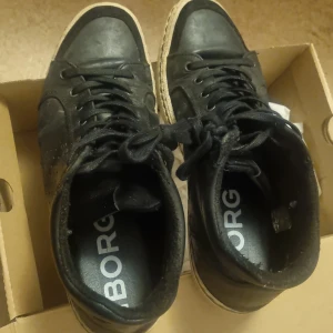 Svarta sneakers från Björn Borg - Snygga svarta sneakers från Björn Borg med högt skaft och klassisk snörning. Skorna har perforerade detaljer på sidorna och vit sula som ger en sportig look. Tillverkade i skinnliknande material och passar perfekt till streetwear-stilen.