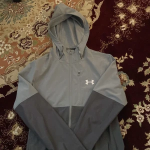 Grå hoodiejacka från Under Armour - Snygg tvåfärgad hoodiejacka från Under Armour i ljusgrått och mörkgrått. Jackan har dragkedja framtill, huva och två sidofickor. Perfekt för träning eller vardagsstil. Under Armour-logga på bröstet. Använd i cirka en och en halv månad. 