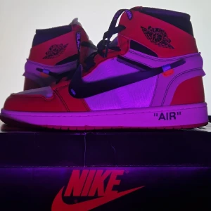 Nike Air Jordan 1 x Off-White Chicago - Nike Air Jordan 1 x Off-White Chicago med ikonisk röd, vit och svart färgkombination. Skorna har tydliga Off-White-detaljer som text på sidan, svarta snören med 'SHOELACES'-print och dekonstruerad look. Tillverkade i skinn och syntetmaterial, med hög siluett och klassisk rund tå.