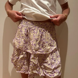 Blommig volangkjol från Americandreams - Supersöt kort volangkjol från Americandreams med blommigt mönster i lila och gul. Kjolen har resår i midjan för en bekväm passform och är perfekt för vår och sommar. 