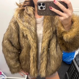 Brun pälsjacka med fluffiga ärmar - Säljer en brun pälsjacka med fluffig look och bred krage. Jackan har långärmade ärmar och är i en loose passform som ger en cool och lyxig vibe. Perfekt för dig som vill sticka ut med en statement piece i garderoben.