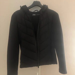 Svart pufferjacka från Zara med huva - Trendig och varm pufferjacka från Zara i storlek S. Jackan har huva, dragkedja framtill och quiltade partier på framsidan, medan ärmarna är släta. Det finns lite sminkfläckar på insidan, men de går lätt bort i tvättmaskin. I övrigt i bra skick och redo för ny användning! 