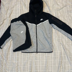 Nike Tech Fleece hoodie svart/grå - Nike Tech Fleece hoodie i svart och grått med vit logga på bröstet. Jackan har hel dragkedja, huva och en snygg kontrast mellan färgerna. Materialet är mjukt och lätt, perfekt för chill eller träning. Ärmarna har en extra ficka med dragkedja för småprylar.