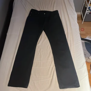 Svarta Levi's 501 jeans W30 L34 - Svarta klassiska Levi's 501 jeans med rak passform. Byxorna har fem fickor, knappgylf och den ikoniska läderpatchen bak i midjan. Tillverkade i slitstarkt jeanstyg och passar perfekt för dig som gillar en tidlös stil.