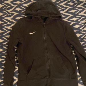 Svart Nike hoodie med dragkedja ( 147-158 barnstorlek) - Svart hoodie från Nike med vit broderad logga på bröstet. Tröjan har huva, hel dragkedja framtill och två stora fickor. Tillverkad i mjuk bomullsblandning, perfekt för chill eller träning. Klassisk och enkel design som funkar till det mesta. Storlek 147-158 barn. Katthår kan förekomma.