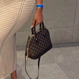  brun handväska - Snygg handväska  i klassiskt brunt och mörkbrunt rutigt mönster. Väskan har svarta skinnhandtag, avtagbar axelrem och guldfärgade metalldetaljer. Perfekt storlek för det viktigaste och ger en lyxig touch till din outfit.