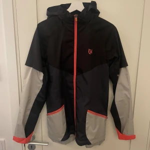 Svart och grå vindjacka från FTKR - Snygg vindjacka från FTKR (footkorner) i svart och grått med röda detaljer. Jackan har huva, hel dragkedja framtill och två fickor med röda dragkedjor. Lätt meshfoder på insidan och cool logga på bröstet. Perfekt för blåsiga dagar och streetstyle.