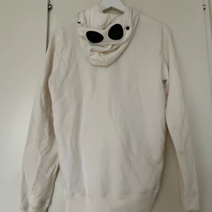 Vit hoodie med goggles från C.P. Company - Vit hoodie från C.P. Company med svarta goggles på huvan. Tror att det är en barnmodell, men passar en S både dam och herr. Tröjan är något missfärgad på vissa partier som runt nacken från användning.