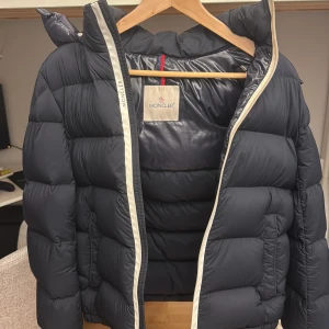 Mörkblå Moncler pufferjacka med huva - Säljer en mörkblå pufferjacka från Moncler med avtagbar huva och klassisk logga på ärmen. Jackan har dragkedja framtill, quiltad design och innerfoder med serietidningsmönster. Perfekt för kalla dagar och riktigt snygg streetstil. 