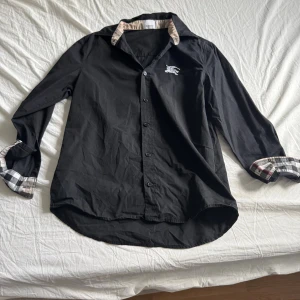 Svart Burberry skjorta med rutiga detaljer - Svart skjorta från Burberry med klassiska rutiga detaljer på insidan av kragen och ärmsluten. Skjortan har knappar framtill, lång ärm och en vit broderad Burberry-logga på bröstet. Tillverkad i bomull och har en snygg, tidlös look.