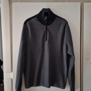 Grå half zip tröja från Hugo Boss - Snygg grå långärmad tröja från Hugo Boss med svarta axelpartier och hög krage med half zip-dragkedja. Tillverkad i en mjuk ullmix som känns lyxig och passar perfekt till lager-på-lager. Enkel och stilren design som funkar till många outfits. Oanvänd men tvättad en gång.