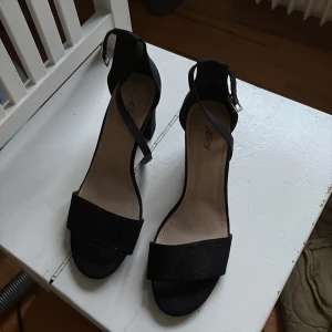 Svarta sandaler med klack och rem - Säljer ett par svarta sandaler från Alley med bred rem över tårna och justerbar ankelrem med spänne. Skorna har en stadig hög klack och är i mockaliknande material. Perfekta för dig som vill ha en stilren och klassisk look.
