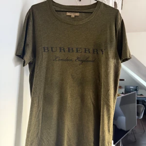 Burberry T-shirt Olivgrön - Olivgrön t-shirt från Burberry med svart logotyp-tryck på bröstet. Klassisk rund halsringning och korta ärmar. Tillverkad i en mjuk blandning av polyester och bomull för en skön känsla. 