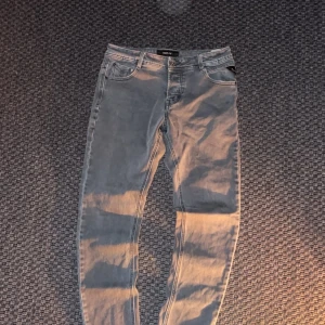 Grå jeans från Replay - Snygga grå jeans från Replay med klassisk femficksdesign och svarta detaljer på knappar och bakficka. Jeansen har en smal passform och är tillverkade i mjukt denimtyg som ger en skön känsla. Perfekta för dig som gillar stilrena och moderna jeans.