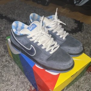 Nike SB Dunk Low - Nike SB Dunk blue lobster med grå mocka och blå detaljer. Swoosh i mörkblått med vit kant, vit snörning och unik blå/vit rutig insida. Skorna har platt sula och rund tå, perfekt för dig som gillar streetwear. Använda 1-2 ggr nyskick.