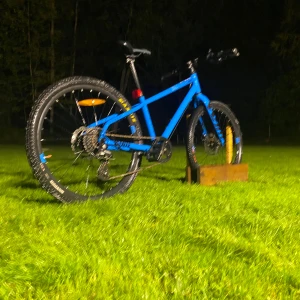 Mountainbike (blå, 27.5 tum) - Snygg blå mountainbike med 27.5-tums hjul, skivbromsar och robust ram. Utrustad med växlar och grova däck för terräng. Cykeln ser ut att vara i gott skick med minimalt slitage, perfekt för både vardag och äventyr. Inga synliga defekter.