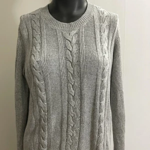 Grå kabelstickad tröja från Lindex - Snygg grå kabelstickad tröja från Lindex med rund hals och långa ärmar. Tröjan har ett klassiskt flätmönster framtill och ribbade muddar vid hals, ärmslut och nederkant. Perfekt för dig som gillar stilrena och mysiga plagg.