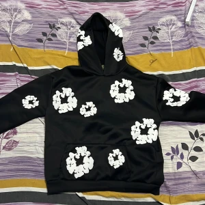 Svart hoodie med vita blommor Denim Tears - Svart hoodie från Denim Tears med stora vita blommor tryckta över hela tröjan, inklusive huvan och fickan. Tröjan har en klassisk känguruficka och huva, samt långa ärmar. Materialet känns mjukt och har en lätt glansig finish, perfekt för dig som gillar statement-plagg.