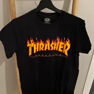 Svart Thrasher t-shirt med flames - Svart t-shirt från Thrasher med ikoniskt flames-tryck i gult och rött på bröstet. Klassisk passform och rund hals. Perfekt för dig som gillar streetwear och skatekultur.
