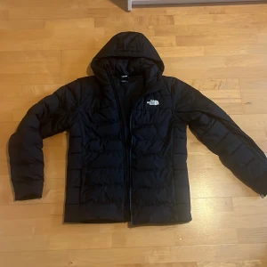 Svart pufferjacka från The North Face - Svart pufferjacka från The North Face med huva och dragkedja. Jackan har en quiltad design och logga på både bröstet och ryggen. Perfekt för kalla dagar och har en clean, sportig look som funkar till det mesta. Passar folk mellan 170-180