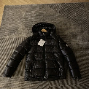 Svart Moncler pufferjacka med huva - Svart pufferjacka från Moncler med glansig finish, huva och dragkedja framtill. Jackan har två fickor med dragkedja och klassisk Moncler-logga på ärmen. Perfekt för kalla dagar och riktigt snygg streetstil.