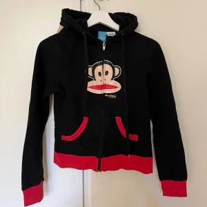 Paul Frank Hoodie med Öron  - Svart hoodie från Paul Frank med Julius-apan tryckt framtill. Tröjan har dragkedja och gullig huva med öron. Kan även mötas upp på Södermalm!