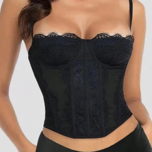 Svart spetskorsett med smala axelband - Säljer en svart korsett med smala axelband och fina spetsdetaljer upptill. Toppen har en figurnära passform och markerad midja, perfekt för att lyfta din outfit. Snyggt mönstrad spets framtill och längs bysten för en extra touch.