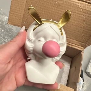 Figur kruka med flicka som blåser bubbla  - Bilderna visar en dekorativ kruka - flicka som blåser bubbla i keramik med guldiga kaninöron och en rosa bubbla. Perfekt för att ljusa upp hemmet med något så ögonfallande figur, man kan även lägga i blommor 🌷 för att göra det mer hem trevligt 🥰😍 höjd: ca: 10 cm 