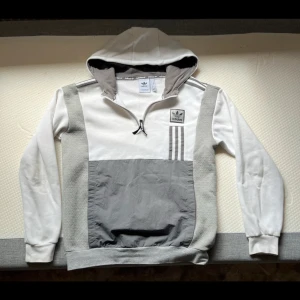 Vit och grå hoodie från Adidas - Snygg hoodie från Adidas i vitt och grått med halv dragkedja och huva. Tröjan har Adidas-logga på bröstet, tre ränder på sidan och stora grå paneler framtill. Mjuk insida och ribbade muddar. Perfekt för en sportig och avslappnad stil.