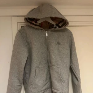 Grå hoodie från Burberry med huva - Säljer en grå hoodie från Burberry med dragkedja och klassiskt rutigt foder i huvan. Tröjan har långa ärmar, fickor framtill och en diskret logga på bröstet. Perfekt för en chill och stilren look.