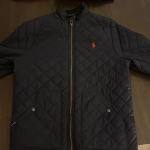 Quiltad jacka från Polo Ralph Lauren - Mörkblå quiltad jacka från Polo Ralph Lauren med röd logga på bröstet. Jackan har dragkedja framtill, två fickor med tryckknappar och en stilren, sportig look. Perfekt för dig som gillar klassisk och clean stil.