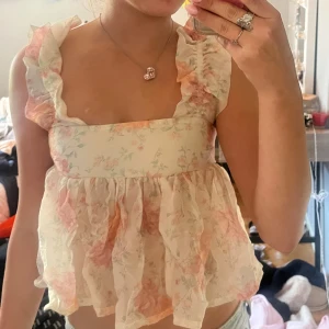 Blommig volangtopp från Divided H&M - Supersöt blommig topp från Divided H&M med volanger på axlarna, fyrkantig ringning och öppen rygg med knyt. Tunt, luftigt material i ljusa pastellfärger med rosa och gröna toner. Perfekt för sommarens alla tillfällen!