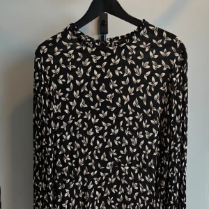 Svart blommig klänning KappAhl - Svart långärmad klänning från KappAhl med vitt blommönster. Klänningen har rund halsringning och dragkedja i ryggen. Materialet är lätt och faller snyggt, perfekt för dig som gillar en stilren men ändå lekfull look.