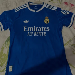 Blå Real Madrid fotbollströja Adidas L - Säljer en blå Real Madrid fotbollströja från Adidas med vita detaljer och klubbmärke på bröstet. Tröjan har klassiska Adidas-ränder på axlarna, vit krage och tryck med Emirates Fly Better. Tillverkad i lätt och ventilerande material.