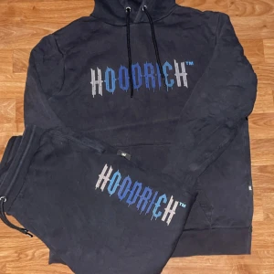 Svart hoodie från Hoodrich med blått tryck - Svart hoodie från Hoodrich med fet blå och vit logga framtill. Klassisk huva med snörning och känguruficka. Mjuk insida och ribbade muddar. Perfekt för dig som gillar streetwear och vill sticka ut med coolt tryck.