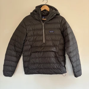 Grå dunjacka från Patagonia med huva - Säljer en grå dunjacka från Patagonia i storlek S. Jackan har huva och en praktisk half zip-dragkedja framtill. Perfekt för kalla dagar och under skidjackan, enkel att styla till det mesta. Klassisk Patagonia-logga på bröstet.