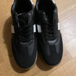 Svarta sneakers från Claudia Ghizzani - Snygga svarta sneakers från Claudia Ghizzani med vit detalj på sidan och bruna gummisulor. Ovandelen är i en mix av skinnimitation och mocka, med svarta skosnören och klassisk rund tå. Perfekta för dig som gillar stilrena och sportiga skor.