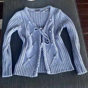 Ljusblå stickad kofta med knyt - Superfin ljusblå stickad kofta från Isolde med flätade detaljer längs v-ringningen och knyt framtill. Koftan har ett snyggt mönster med små bollar och långa ärmar. Perfekt att slänga över en topp för en chill look. Endast swish betalning med spårbart frakt 59 kr
