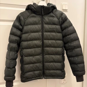 Mörk grön dunjacka Peak Performance 170 - Snygg mörk grön dunjacka från Peak Performance med huva och dragkedja framtill. Jackan har quiltad design, ribbade muddar och två fickor med dragkedja. Perfekt för kalla dagar och riktigt skön att bära.