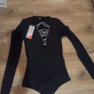 Svart långärmad body med snörning - Svart långärmad body från New Yorker med snygg silverfärgad snörning framtill i urringningen. Kroppsnära passform och djup v-ringning som ger en cool och trendig look. Perfekt att styla med jeans eller kjol för en edgy vibe.