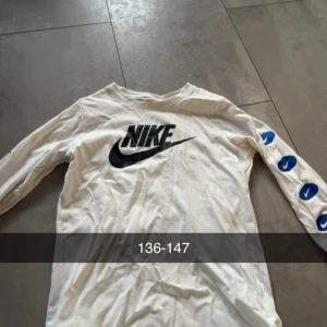 Vit långärmad Nike-tröja - Vit långärmad tröja från Nike med stor svart logga på bröstet och blå Nike-symboler längs ena ärmen. Perfekt för dig som gillar sportig och clean stil. Tillverkad i mjuk bomull för skön känsla hela dagen.