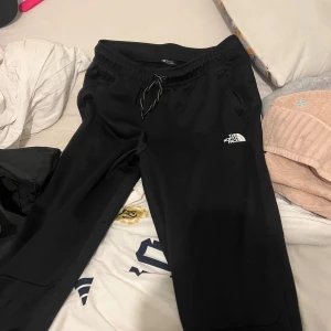 Svarta mjukisbyxor från The North Face - Svarta mjukisbyxor från The North Face med dragsko i midjan och logga på låret. Byxorna har en avslappnad passform och är perfekta för chill eller träning. Mjukt material och elastisk midja för extra komfort.