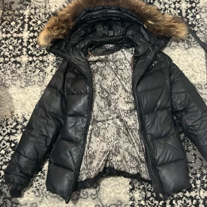 Svart pufferjacka Hollies med fuskpäls - Snygg svart pufferjacka från Hollies med stor huva och fuskpäls. Jackan har dragkedja, bälte i midjan och långa ärmar. Insidan har ett unikt blommigt mönster. Perfekt för kalla dagar när du vill vara både varm och stilren.