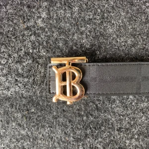 Burberry vändbart bälte med guldspänne - Snyggt vändbart bälte från Burberry med klassiskt rutmönster på ena sidan i beige, rött och grått, och diskret svart rutmönster på andra sidan. Spännet är i guld med Burberrys ikoniska TB-logo. Perfekt accessoar för att lyfta din outfit.