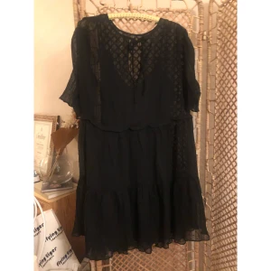 Svart volangklänning från H&M - Svart klänning från H&M i storlek 46 med volangdetaljer och transparenta partier. Klänningen har kort ärm, knytning vid halsen och ett diskret mönster i tyget. Luftig och flowy modell som ger en avslappnad men ändå chic look.