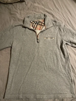Grå half zip tröja  - Snygg grå tröja från Burberry med klassiskt rutigt foder i kragen och broderad logga på bröstet. Tröjan har half zip-dragkedja och lång ärm, perfekt för lager-på-lager. Mjuk bomullskänsla och stilren design.